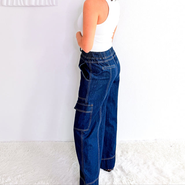A woman wearing a white tank top and dark blue wide-leg cargo jeans stands in profile against a white wall, looking back over her shoulder. A textured white wall art piece hangs on the left, and a white rug covers the floor.