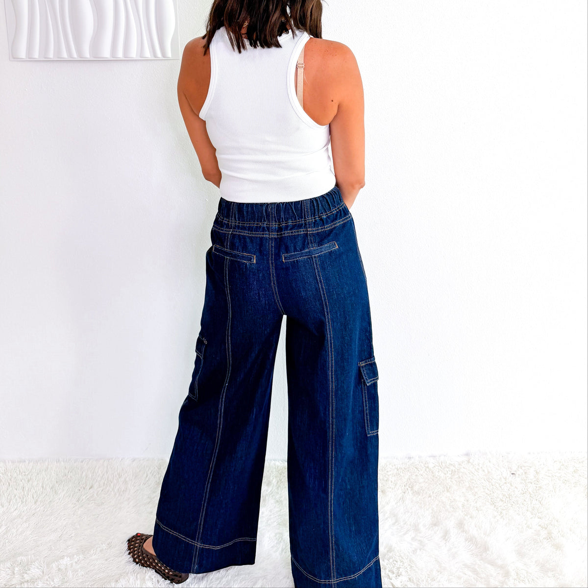 A woman viewed from behind wearing a white tank top and dark blue wide-leg cargo pants, standing in a bright room with a white wall and textured rug.