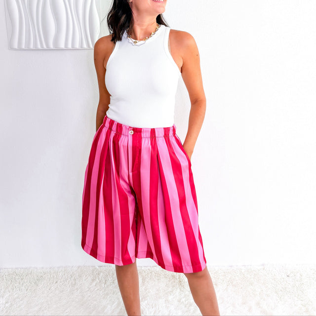 A woman wearing a white sleeveless top and bright pink and red vertically striped high-waisted shorts, standing against a white wall with a textured white panel.