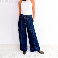 A woman wearing dark blue wide-leg denim cargo pants, a white sleeveless top, and sunglasses, standing against a white wall with a textured white rug.