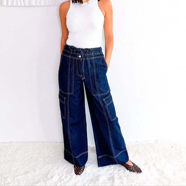 A woman wearing dark blue wide-leg denim cargo pants, a white sleeveless top, and sunglasses, standing against a white wall with a textured white rug.