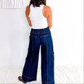 A woman viewed from behind wearing a white tank top and dark blue wide-leg cargo pants, standing in a bright room with a white wall and textured rug.
